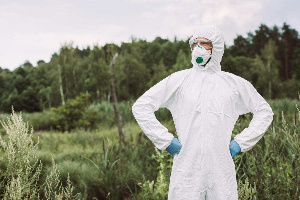 confident male scientist in protective mask, googles and suit standing with hands on waist in meadow - Photo, Image