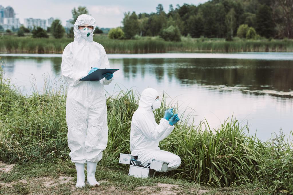 selektiver Fokus einer Wissenschaftlerin in Schutzmaske und Anzug, die in Klemmbrett schreibt, während ihr Kollege Wasserproben in Flussnähe im Freien abgibt  - Foto, Bild