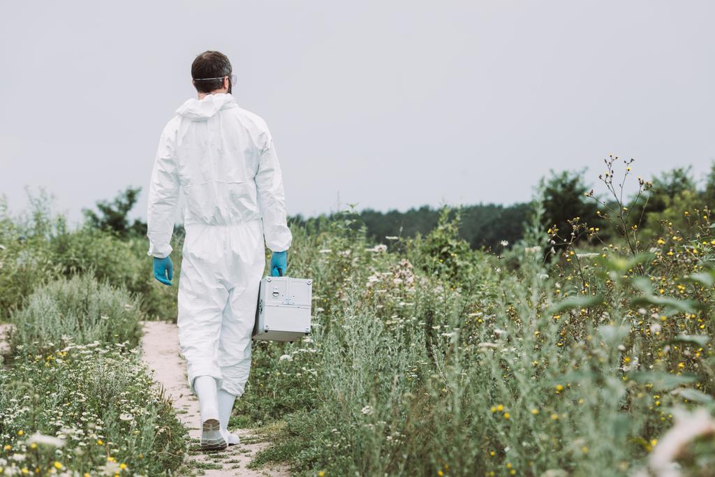 vista trasera del científico masculino en traje de protección caminando con maleta de trabajo en el prado
  - Foto, Imagen