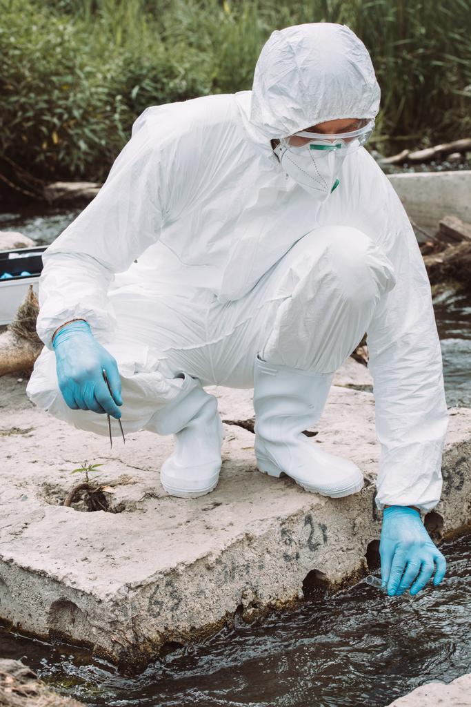 male scientist in protective suit and mask taking sample of water in test flask outdoors  - Photo, Image