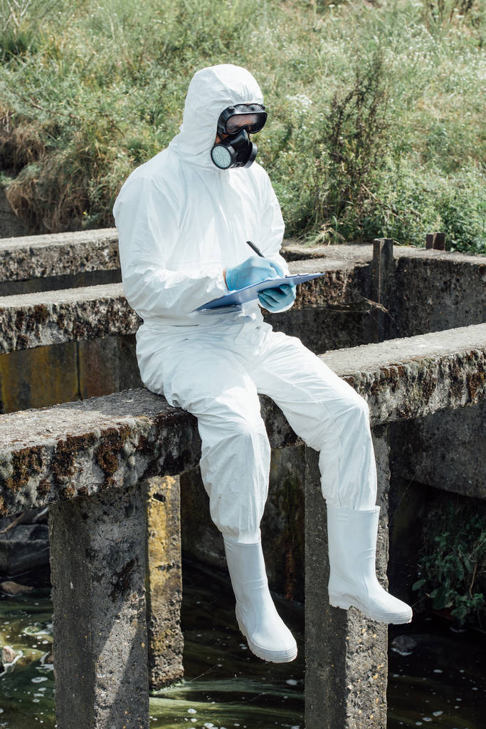 selective focus of male scientist in protective mask and suit writing in clipboard while sitting near sewerage  - Photo, Image