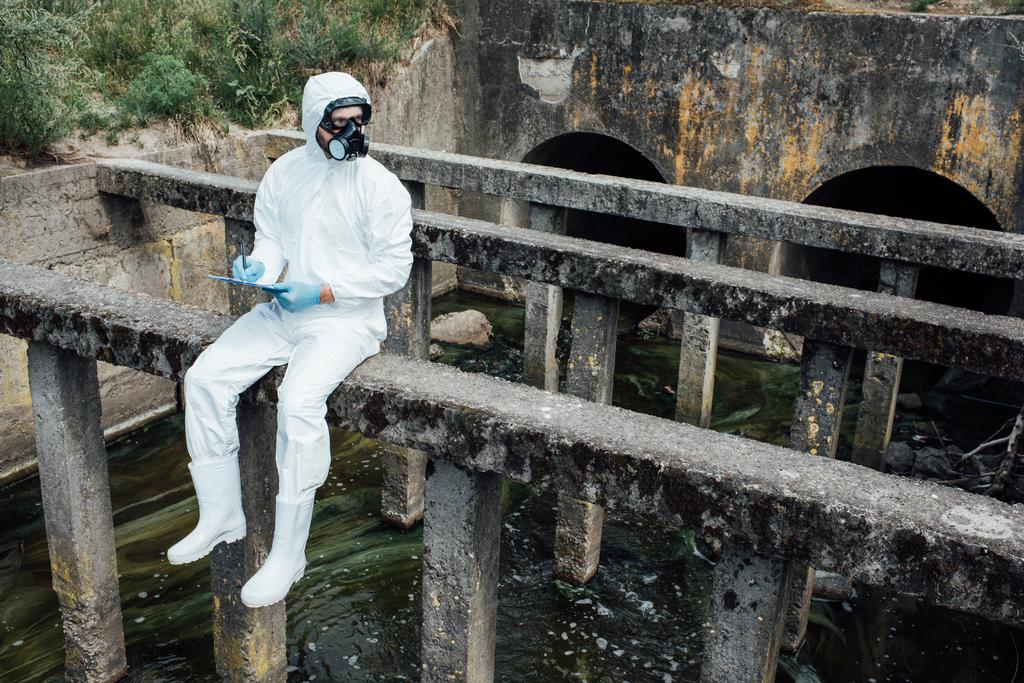 männliche Wissenschaftler in Schutzmaske und Anzug schreiben in Klemmbrett, während sie in der Nähe der Kanalisation sitzen  - Foto, Bild