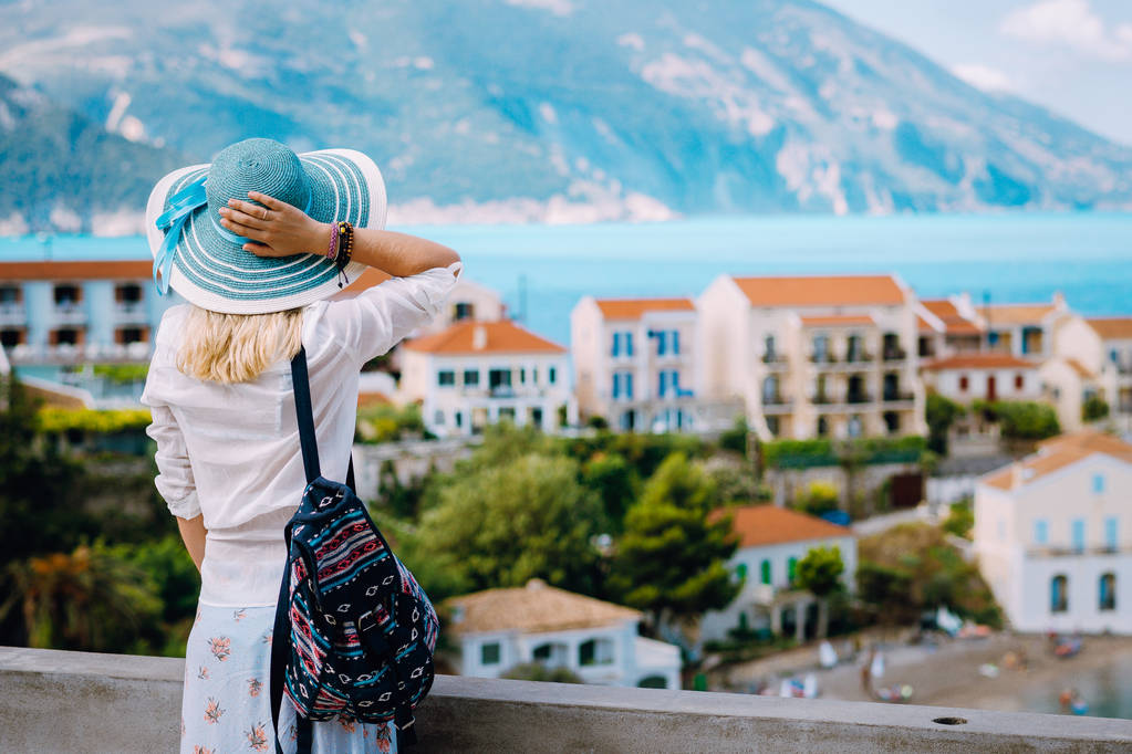 Kobieta turysta podziwiając widok kolorowe spokojnej miejscowości Assos na rano. Młody stylowy model kobiet na sobie niebieski Kapelusz przeciwsłoneczny i białe ubrania korzystających lato wakacje w Kefalonia, Grecja - Zdjęcie, obraz