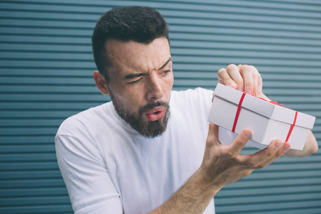 Suspicious man is trying to open box to see what is there. He doesnt want to wrap ribbons. Isolated on striped and blue background. - Photo, Image