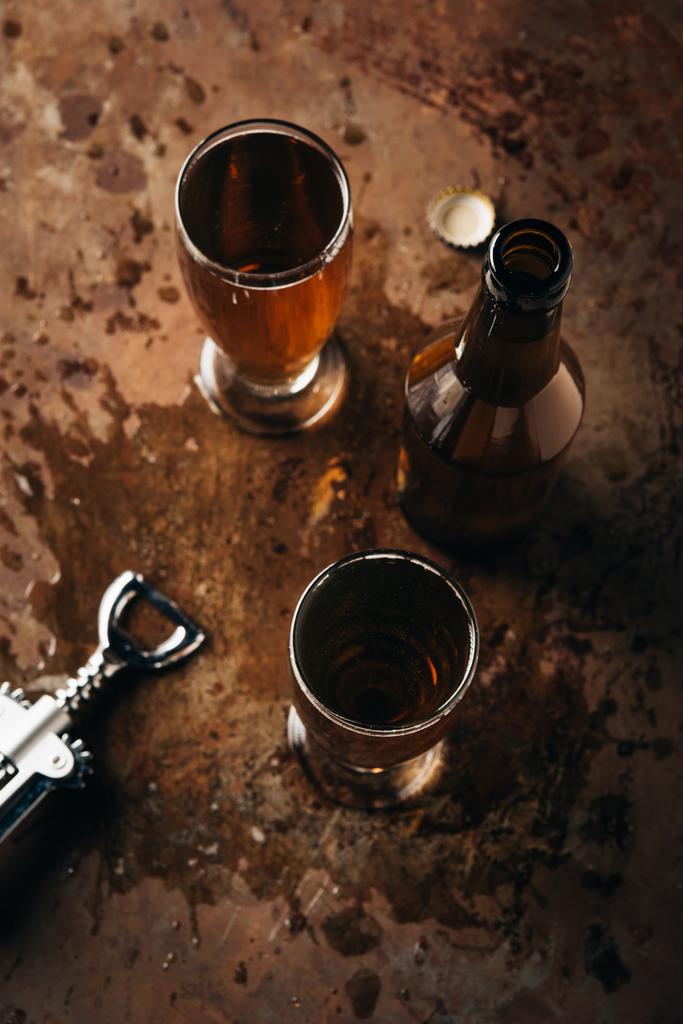 bovenaanzicht van mokken van bier, fles en fles opener op roest oppervlak - Foto, afbeelding