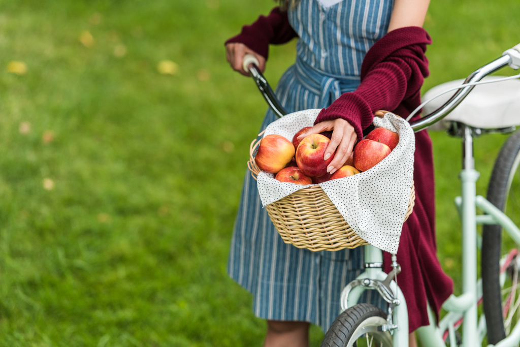 обрезанный вид девушки с плетеной корзиной, полной свежих яблок на велосипеде
 - Фото, изображение