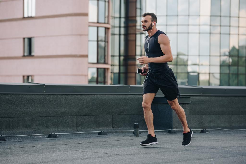boční pohled pohledný sportovec běh a poslech hudby s smartphone na střeše - Fotografie, Obrázek