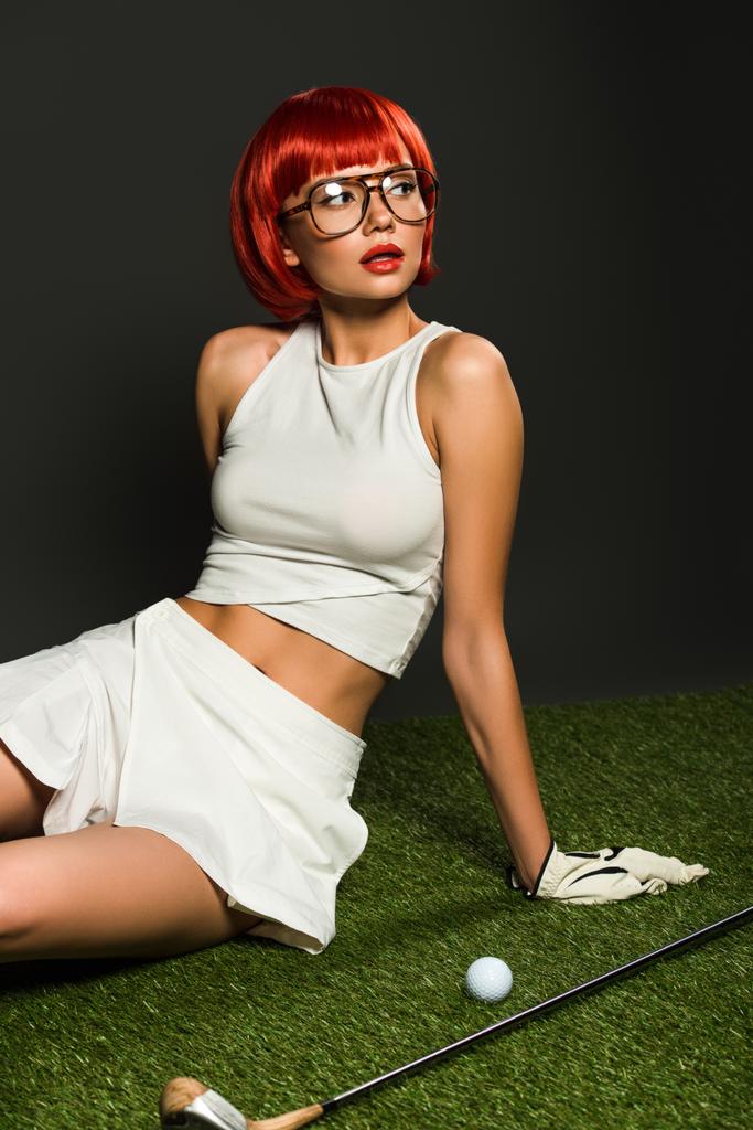 attractive young woman with red bob cut sitting on green grass with golf equipment on grey - Photo, Image