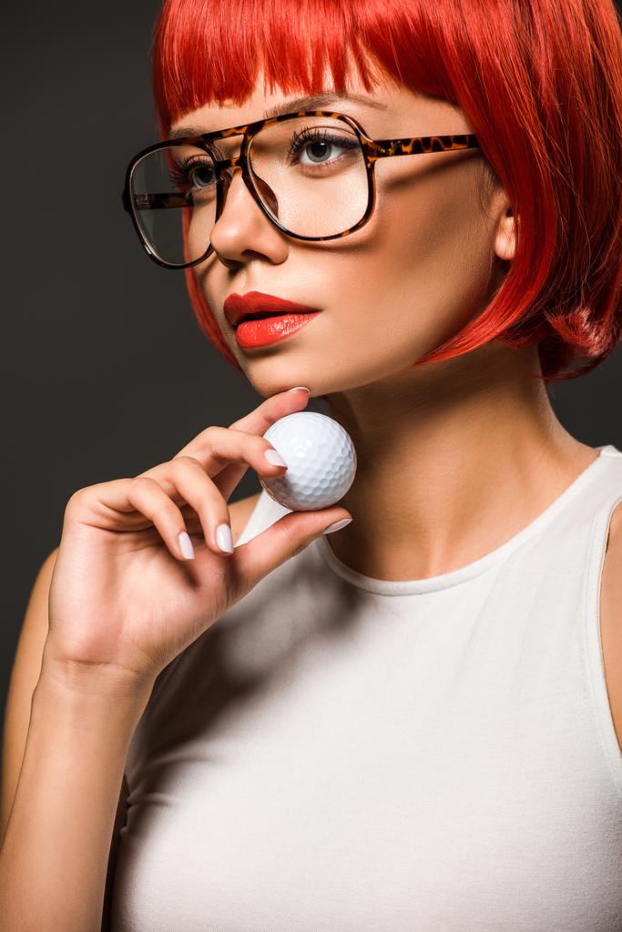 close-up portrait of attractive young woman with red bob cut and stylish eyeglasses holding golf ball isolated on grey - Photo, Image