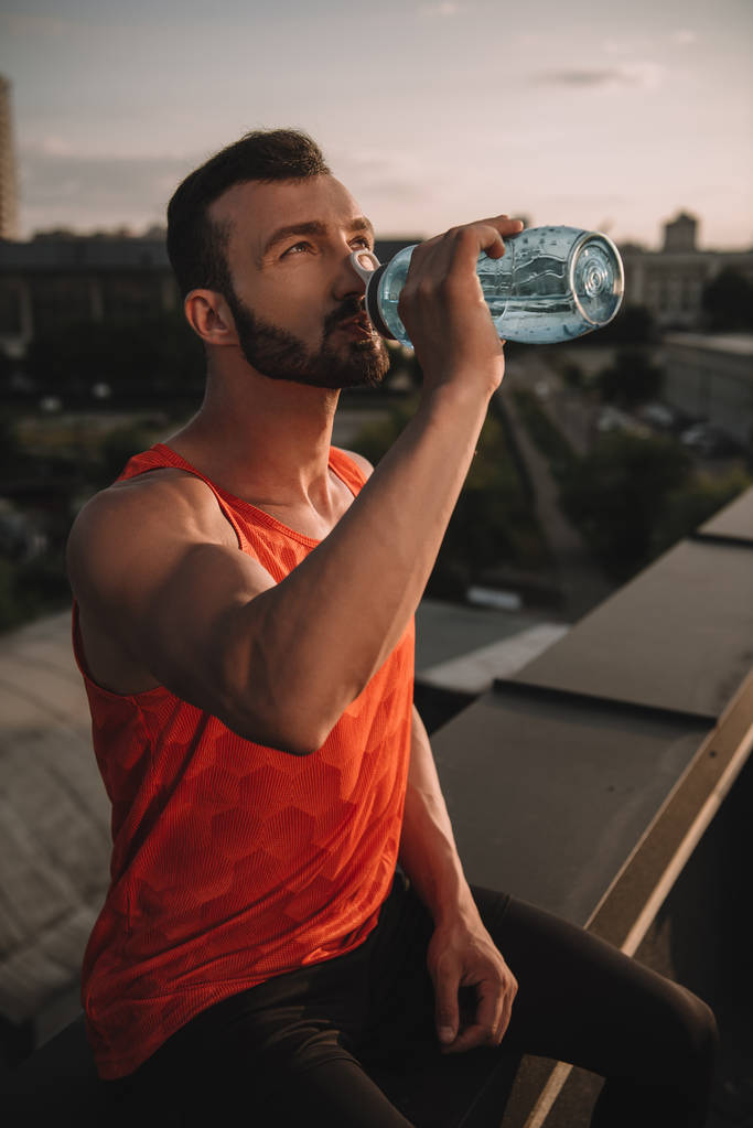 handsome sportsman drinking water from sport bottle on roof - Photo, Image