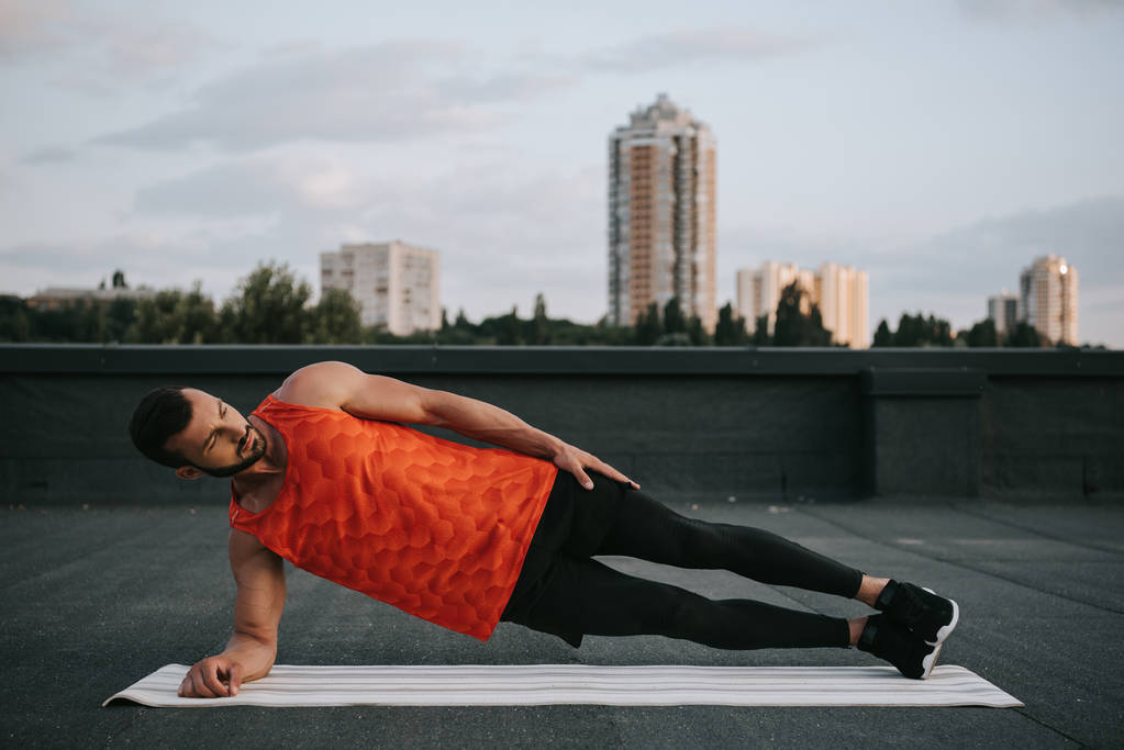 красивый спортсмен делает боковую доску на коврике для йоги на крыше
 - Фото, изображение