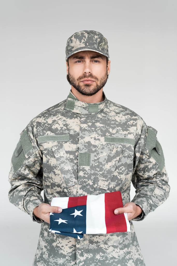 Porträt eines Soldaten in Militäruniform mit amerikanischer Flagge in den Händen vor grauem Hintergrund - Foto, Bild