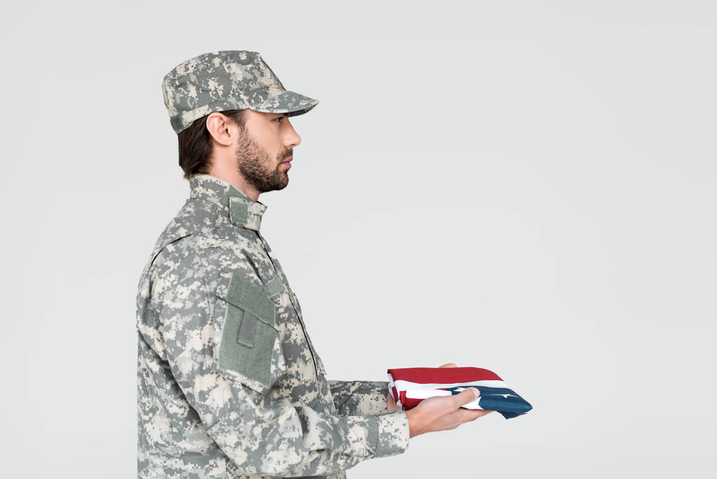 Zijaanzicht van mannelijke soldaat in militair uniform met de Amerikaanse vlag in handen op grijze achtergrond - Foto, afbeelding