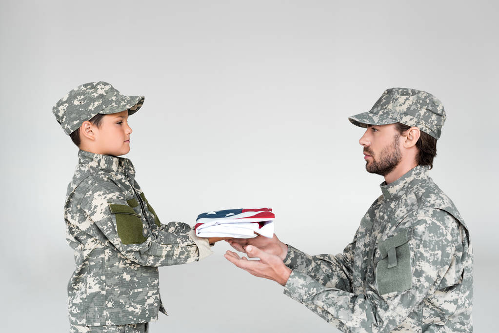 vista lateral do menino em roupas de camuflagem dando dobrado bandeira americana para soldado no fundo cinza
 - Foto, Imagem