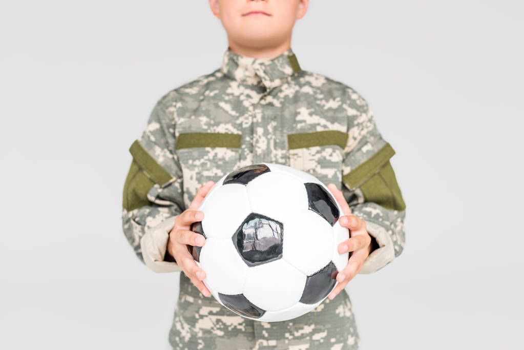 gedeeltelijke weergave voor kid in militair uniform met voetbal in handen geïsoleerd op grijs - Foto, afbeelding