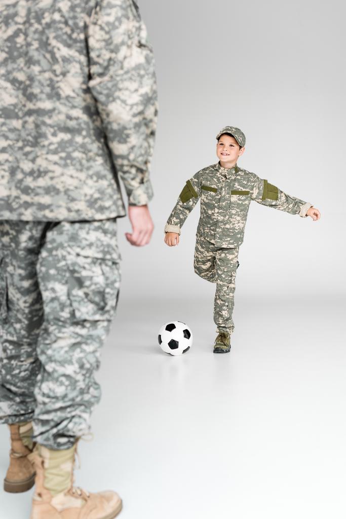 partial view of father and son in military uniforms playing soccer on grey background - Photo, Image