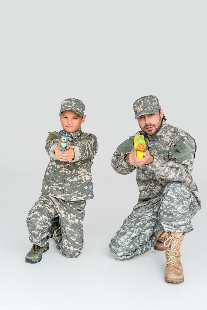 Vater und Sohn in Militäruniformen mit Spielzeug-Wasserpistolen auf grauem Hintergrund - Foto, Bild