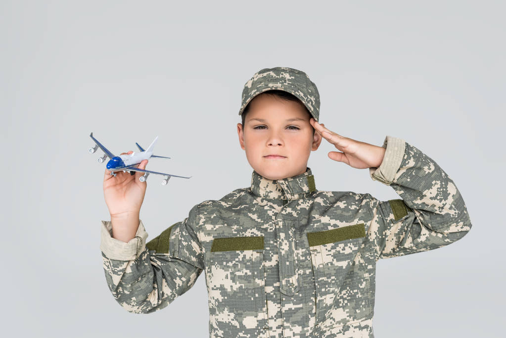 portrait of little boy in military uniform with toy plane in hand saluting isolated on grey - Photo, Image