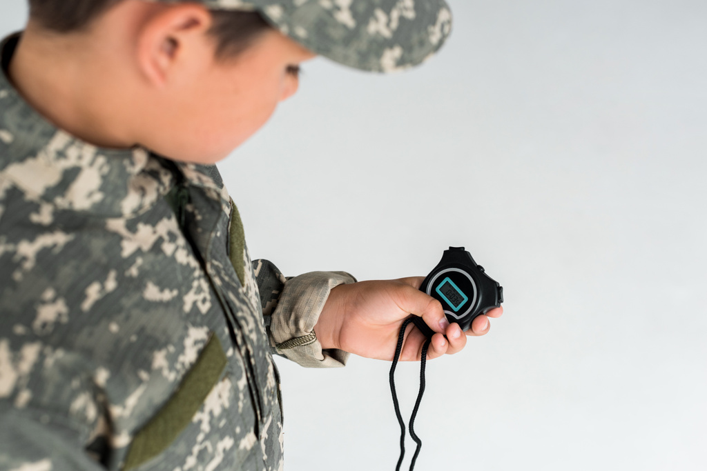 partial view of kid in military uniform looking at stopwatch in hand on grey background - Photo, Image