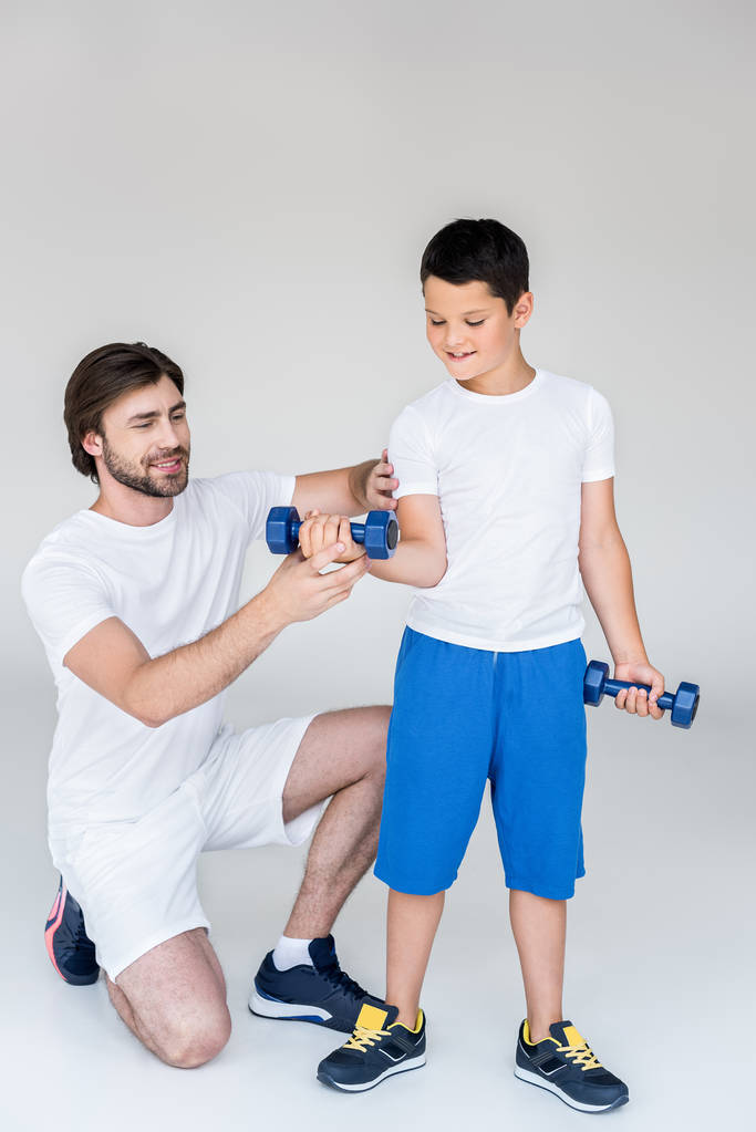 father helping little son with dumbbells to exercise on grey background - Photo, Image