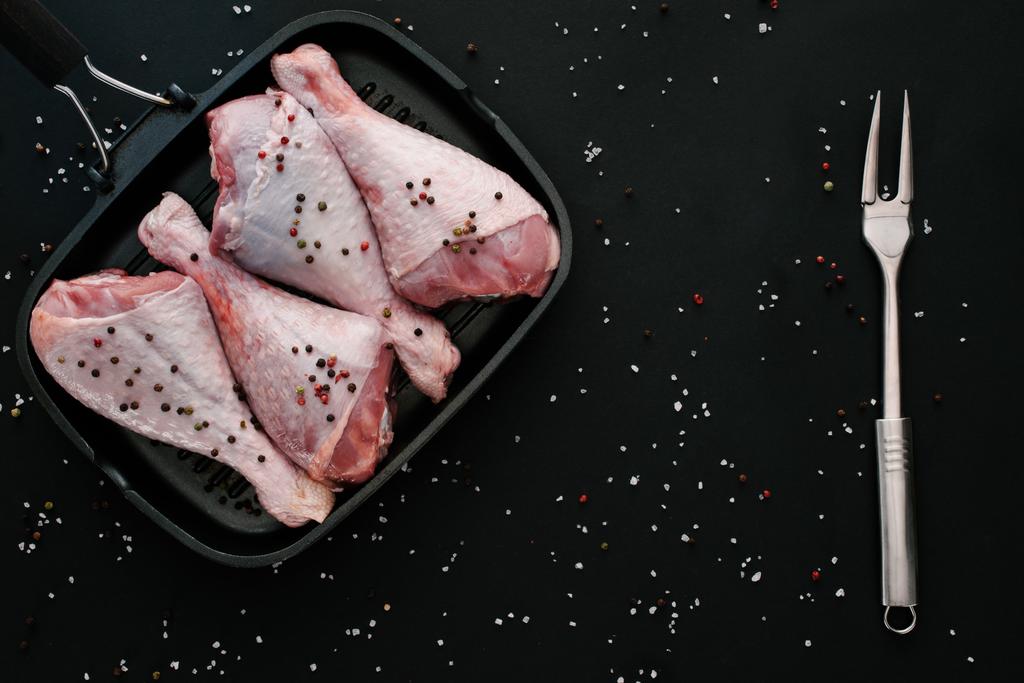 top view of raw chicken legs with pepper corns and salt in grill pan with meat fork on black - Photo, Image