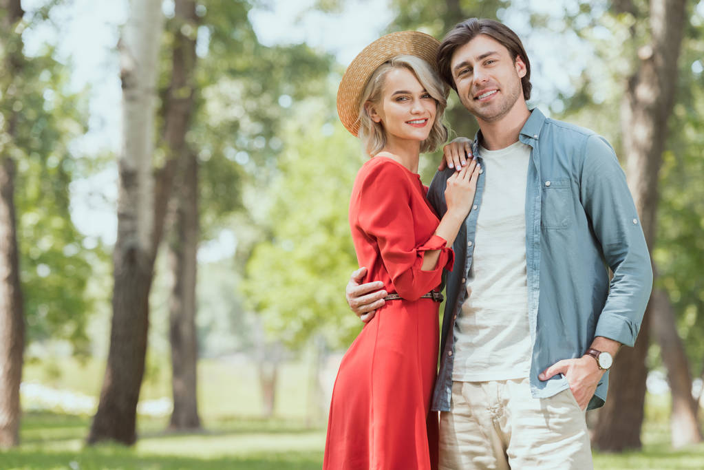 novia sonriente y novio abrazándose en el parque y mirando a la cámara
 - Foto, Imagen