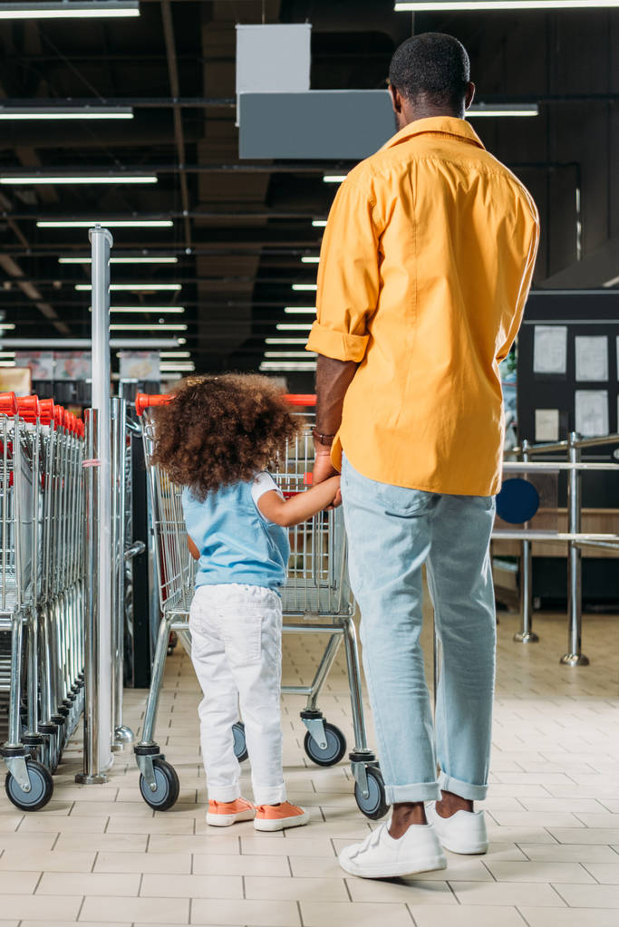 Rückansicht eines afrikanisch-amerikanischen Mannes, der mit seiner Tochter in der Nähe eines Einkaufswagens in einem Verbrauchermarkt steht - Foto, Bild