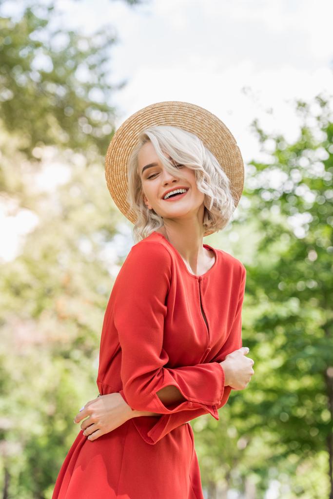 χαμογελώντας ελκυστικό κορίτσι με κόκκινο φόρεμα και ψάθινο καπέλο στο πάρκο - Φωτογραφία, εικόνα