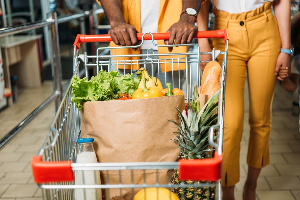 abgeschnittenes Bild von jungem Paar, das Einkaufswagen mit Lebensmitteln im Supermarkt trägt  - Foto, Bild