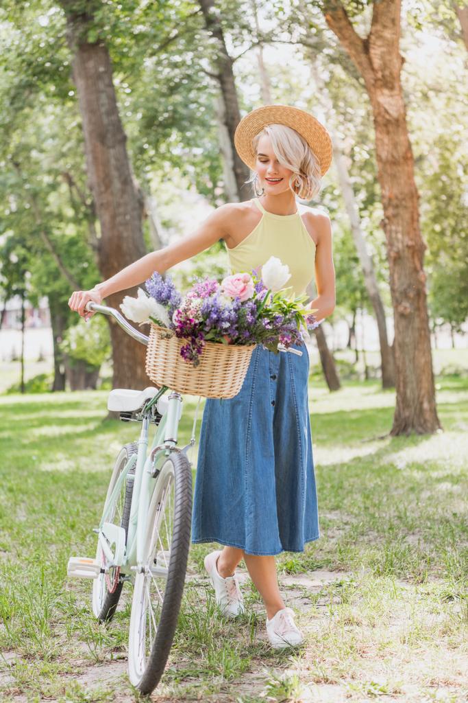 Piękne blond dziewczyna spaceru z rowerem z koszem kwiatów w parku - Zdjęcie, obraz