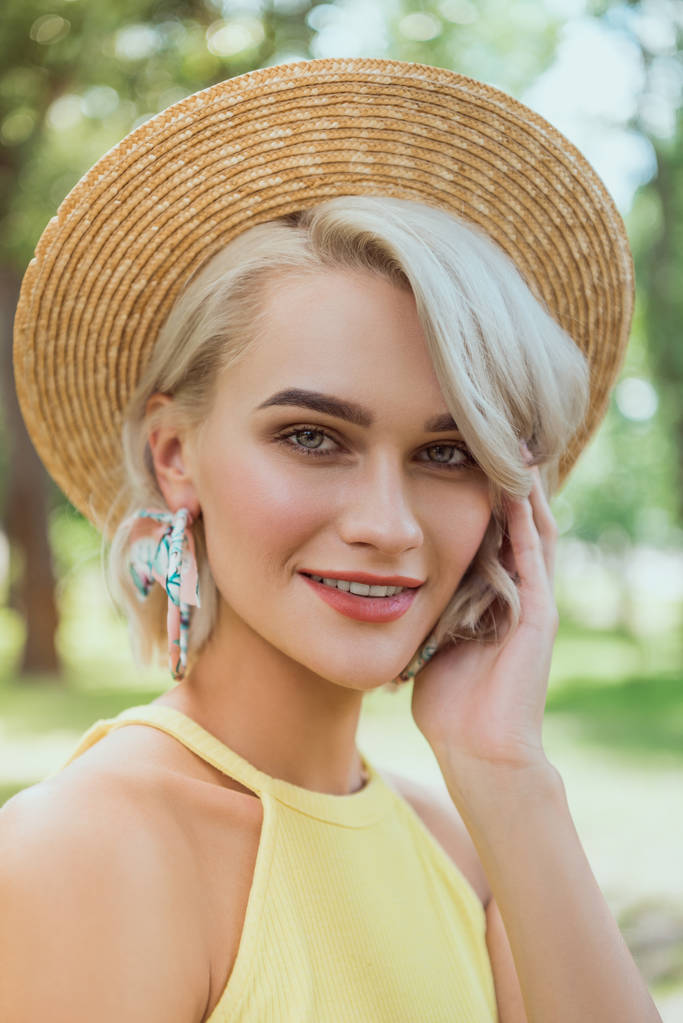 Porträt eines attraktiven blonden Mädchens mit Strohhut, das im Park in die Kamera blickt - Foto, Bild