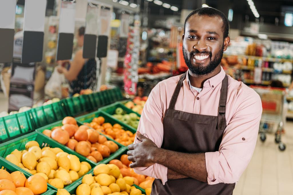 assistente negozio afroamericano maschio in grembiule in piedi con braccia incrociate vicino reparto frutta in ipermercato
  - Foto, immagini