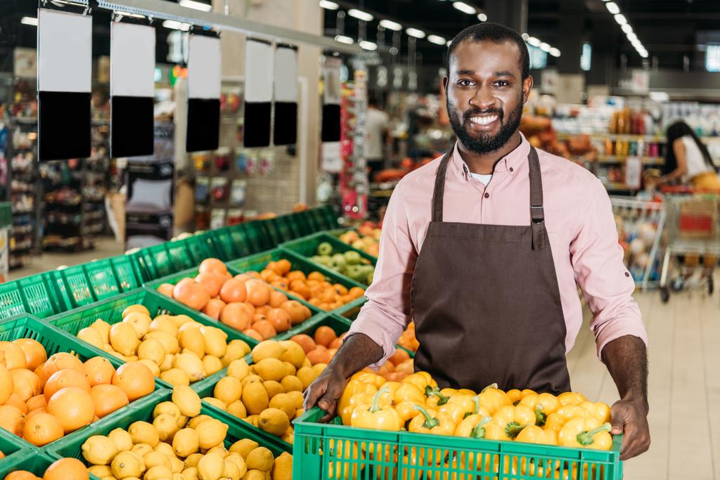 happy african american male shop assistant in apron holding box with bell peppers in grocery store - Photo, Image
