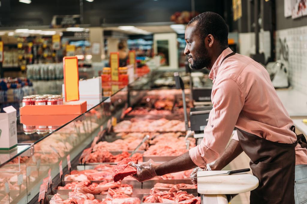 puoli näkymä afrikkalainen amerikkalainen uros teurastaja esiliina antaa pihvi raakaa lihaa supermarketissa
  - Valokuva, kuva