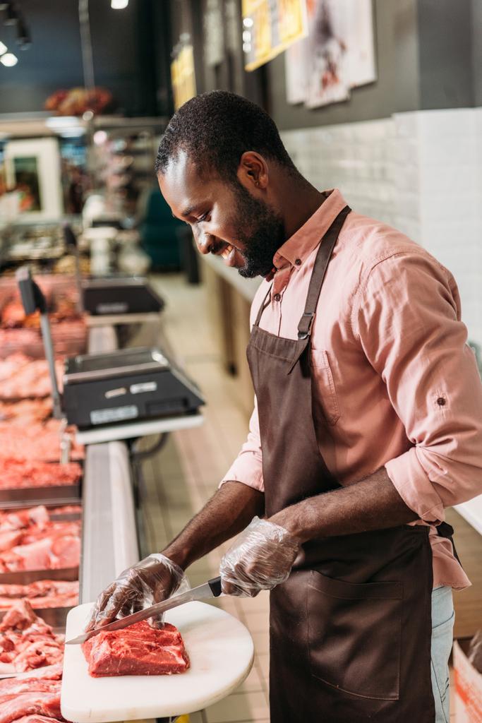 sonriente afroamericano asistente de tienda masculina en delantal corte de filete de carne cruda en el supermercado
  - Foto, imagen