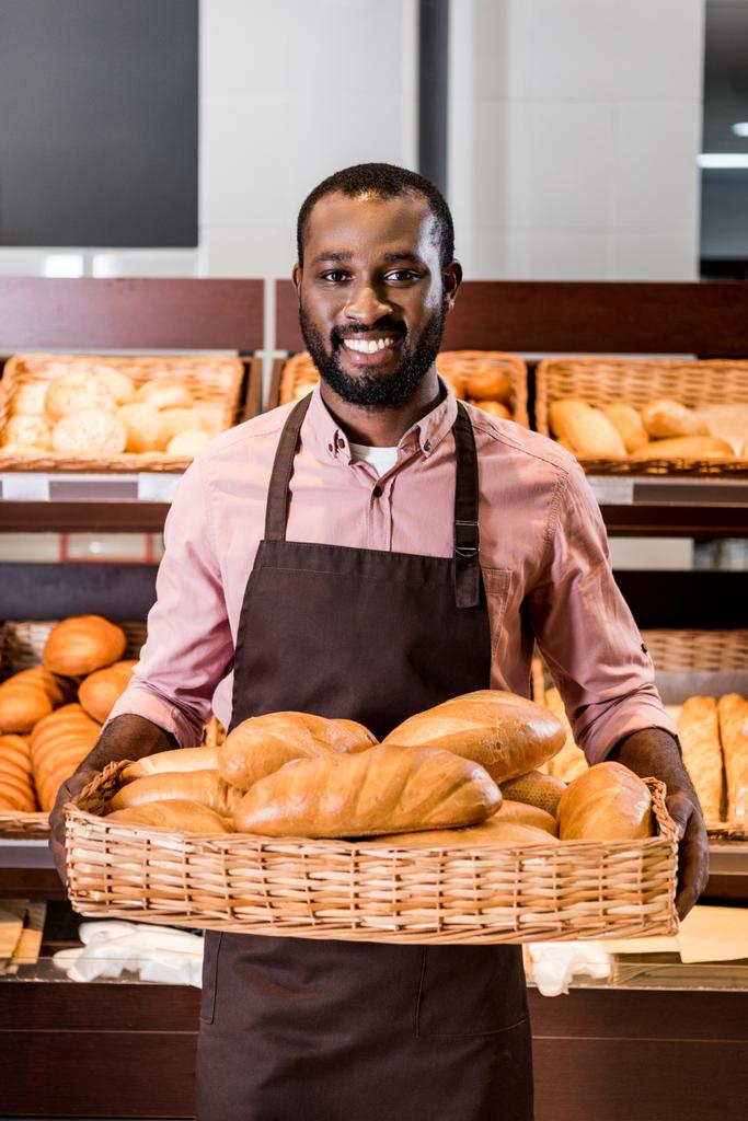 smiling african american male shop assistant in apron holding with loaves of bread in supermarket  - Photo, Image