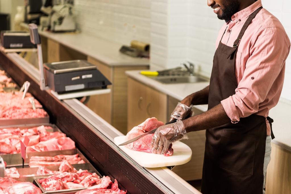 Μερική άποψη της χαμογελώντας αφροαμερικάνων άνδρες κατάστημα βοηθός στην ποδιά μπριζόλα κοπής από το ωμό κρέας στην υπεραγορά - Φωτογραφία, εικόνα