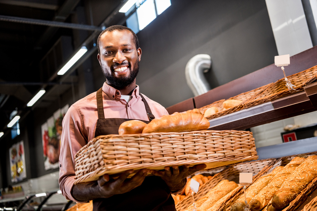 selectieve aandacht van lachende mannelijke Afrikaanse Amerikaanse winkelbediende in schort holding broden in supermarkt - Foto, afbeelding