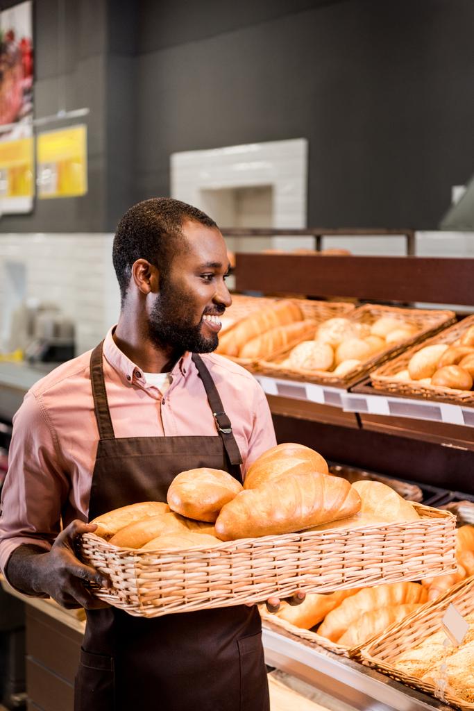 africano americano tienda asistente en delantal celebración de panes en el supermercado
  - Foto, Imagen