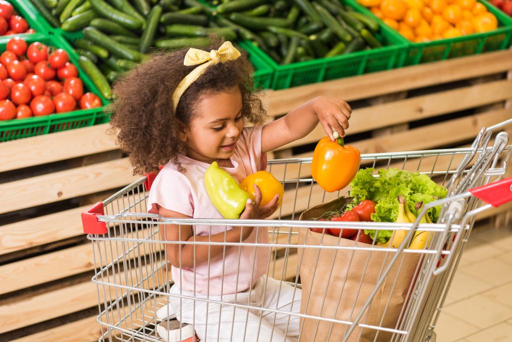 niño afroamericano sonriente poniendo pimientos en bolsa de papel mientras está sentado en el carrito de la compra en el supermercado
  - Foto, imagen