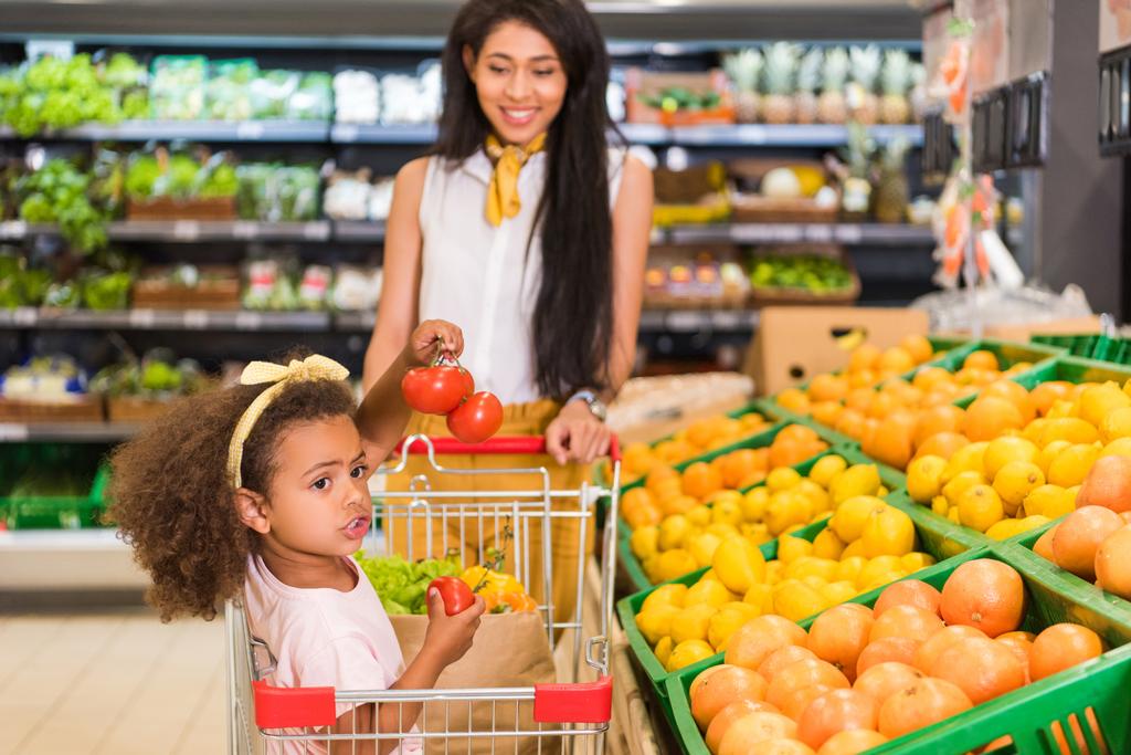 Afro-Amerikaanse kindje zit in het winkelwagentje terwijl haar moeder dragen in de buurt van fruit afdeling in supermarkt  - Foto, afbeelding