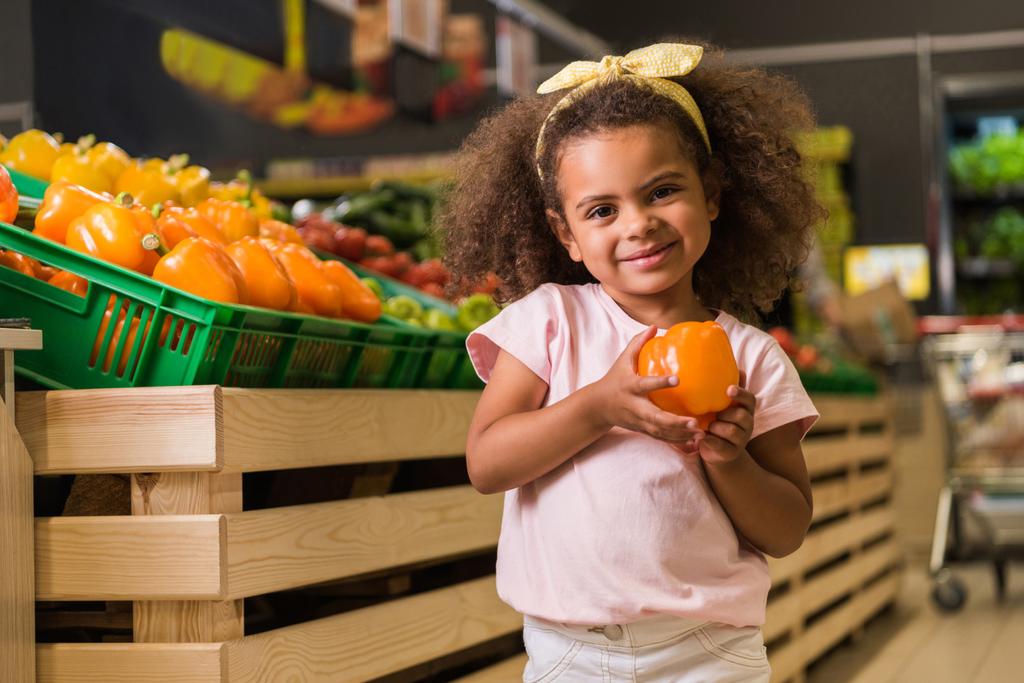 selektiver Fokus eines entzückenden afrikanisch-amerikanischen Kindes, das mit Paprika im Supermarkt steht und in die Kamera blickt - Foto, Bild