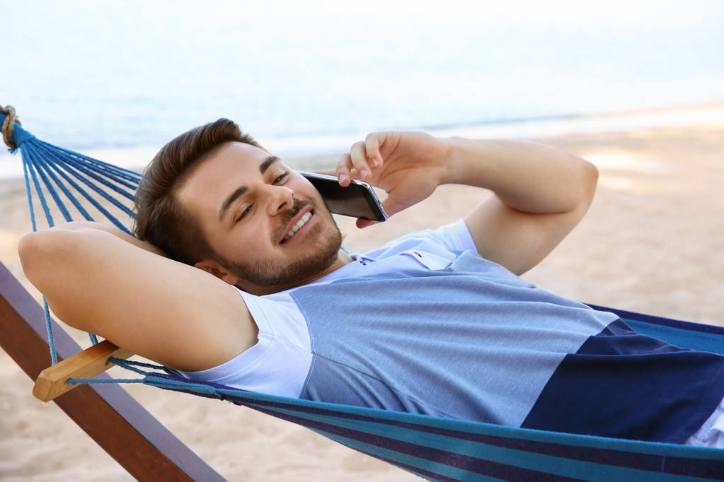 A fiatalember beszél telefonon függőágyban a tengerparton - Fotó, kép