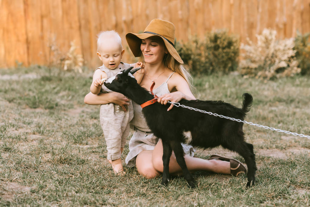 усміхнена мати в капелюсі і маленький син мацає домашню козу на зеленій траві в сільській місцевості
 - Фото, зображення