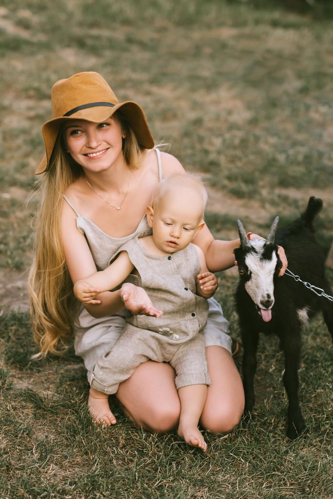 smiling mother in hat, little son and domestic goat on green grass at countryside - Photo, Image