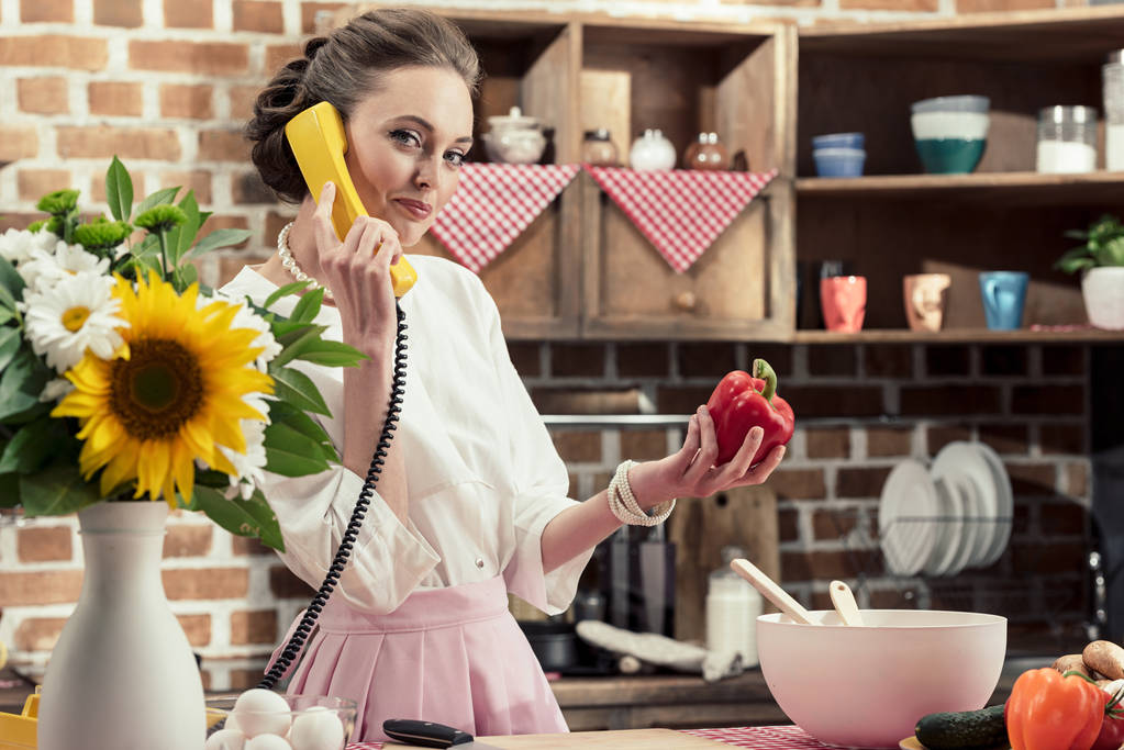 glimlachend volwassen huisvrouw door retro draadloze telefoon praten en kijken naar camera op keuken - Foto, afbeelding