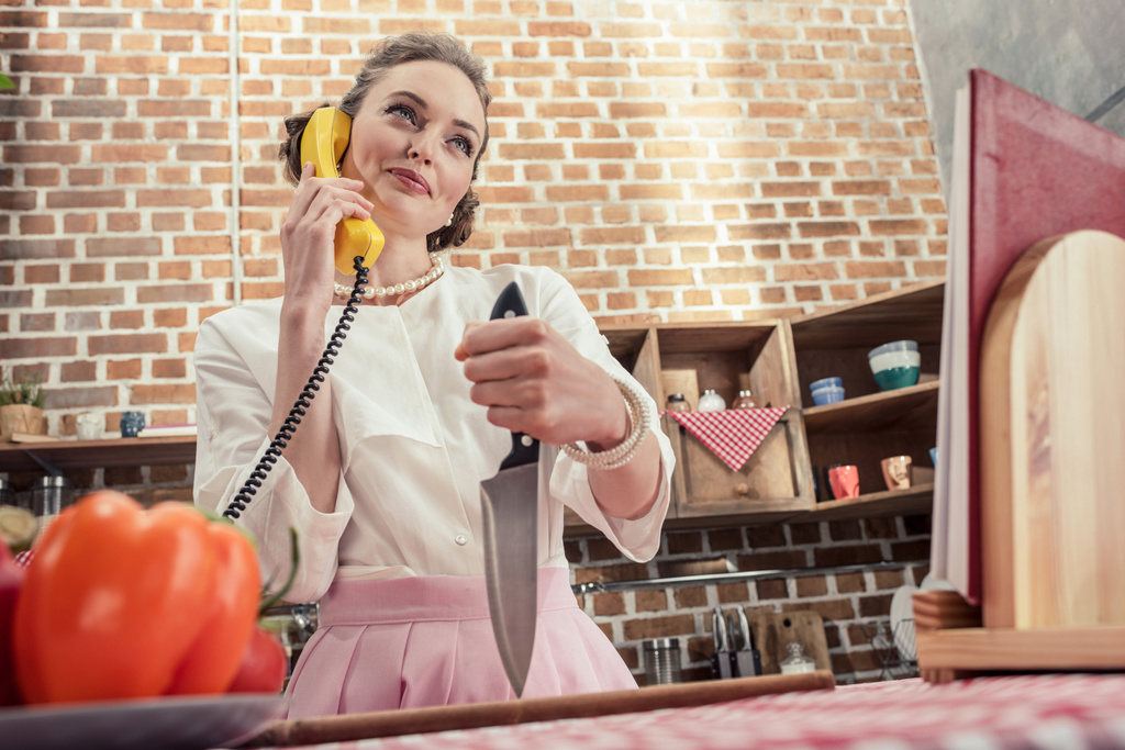 pohja näkymä hymyilevä aikuinen kotiäiti veitsi puhuu keltainen langallinen puhelin keittiössä
 - Valokuva, kuva