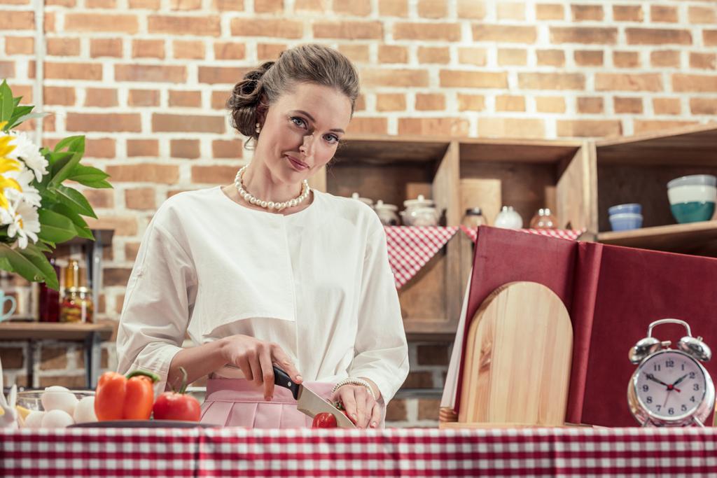 sorrindo adulto dona de casa em roupas vintage cortando tomate e olhando para a câmera na cozinha
 - Foto, Imagem