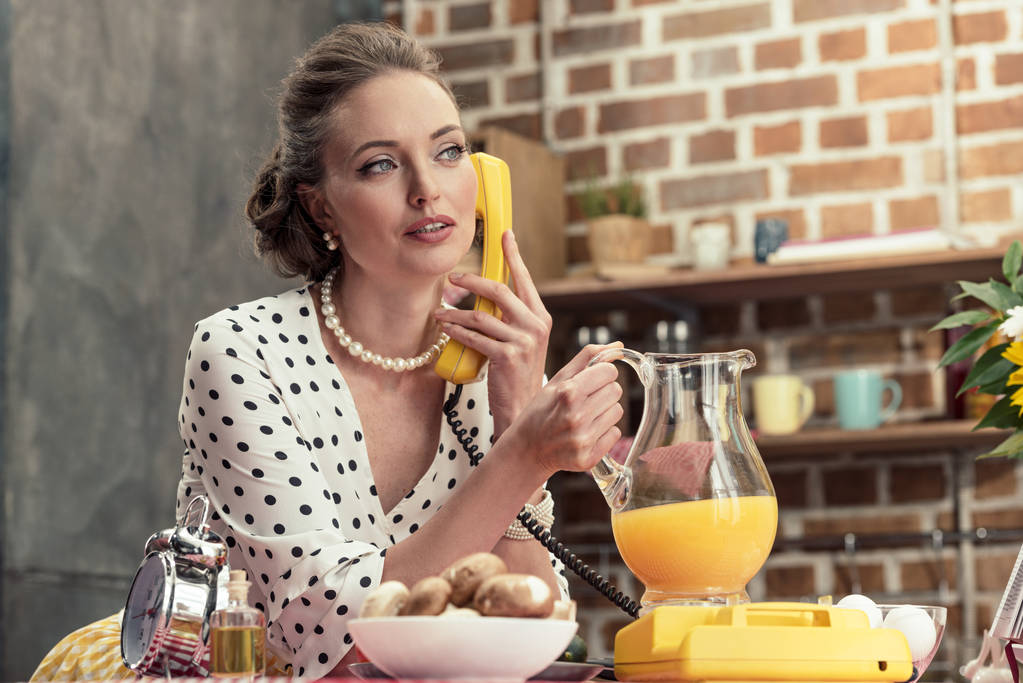 attractive adult housewife holding jug of orange juice talking by vintage wired phone at kitchen - Photo, Image