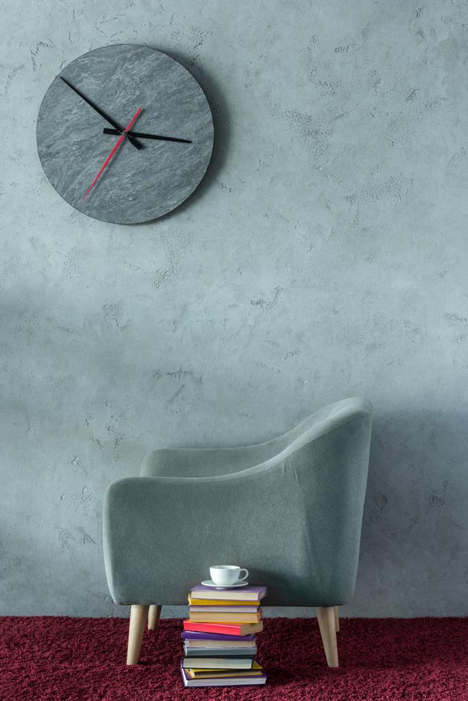 stack of books and cup of coffee near grey armchair in office, clock on wall - Photo, Image